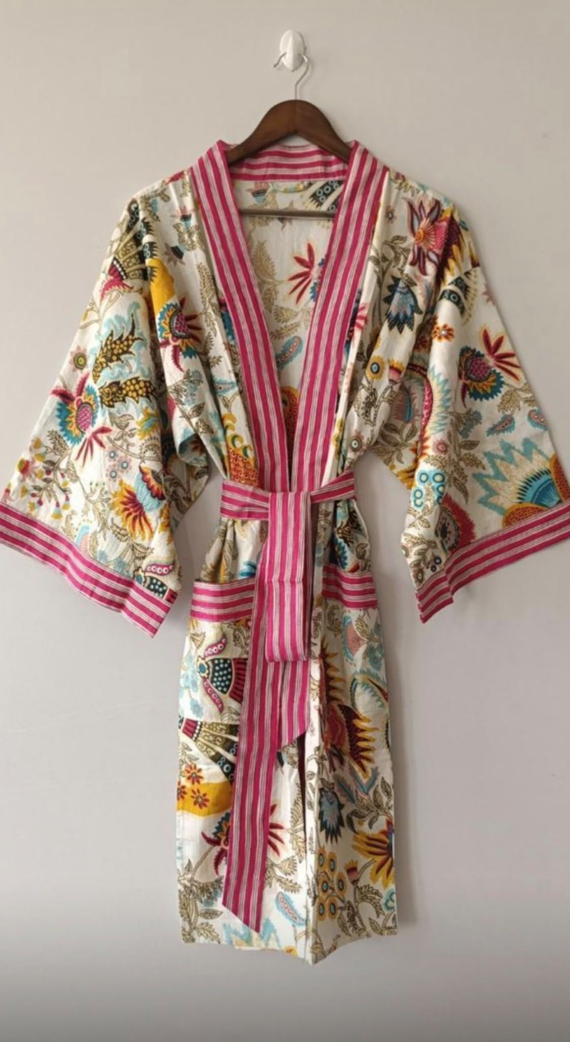 A kimono-style robe hanging on a wooden hanger against a plain light gray wall. The robe has a multicolor floral and bird print on a cream background with pink and white vertical stripes on the collar, cuffs, and waist tie belt.
