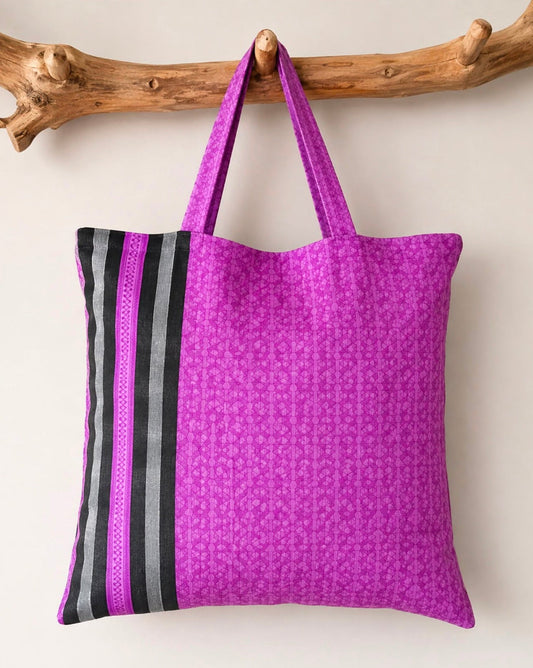 A purple patterned fabric tote bag with black and gray striped side panels and matching purple shoulder straps, hanging from a natural wood branch against a plain light background.
