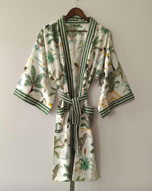 A white cotton bathrobe with a green and multicolor jungle print, featuring green striped trim and a matching tie belt, hanging on a wooden hanger against a plain light gray wall.
